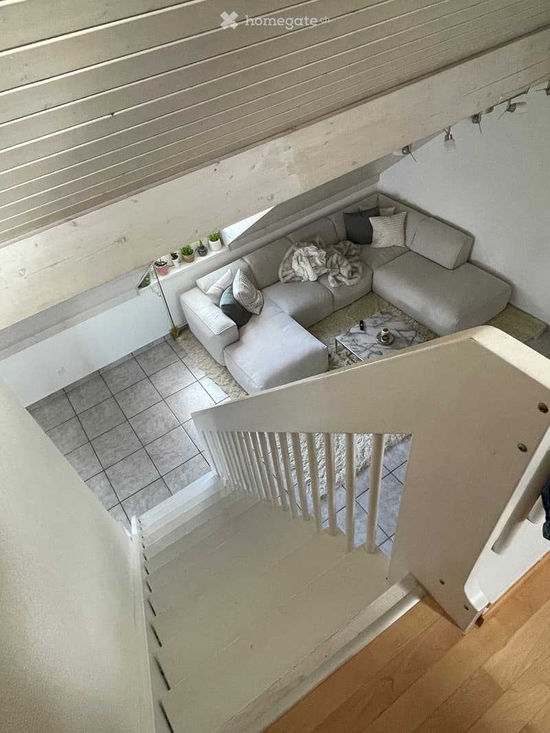 Modern living room with a grey sectional sofa, coffee table, small side table, and decorative items on a shelf. The room features a staircase leading to another level.