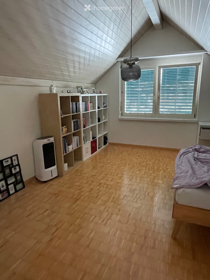 Spacious attic bedroom with wooden flooring, large windows, a bed with a purple blanket, a bookshelf filled with books, an air purifier, and a hanging lamp.