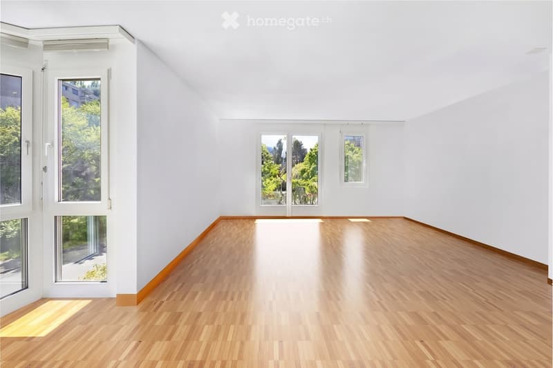 Empty room with white walls, wooden flooring, and multiple windows offering natural light and a view of greenery outside.