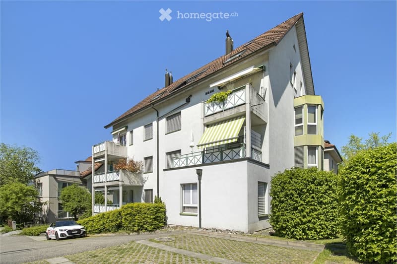 A multi-story residential building with white walls, green accents, and multiple balconies. The building features a tiled roof, large windows, and is surrounded by greenery and a paved driveway.