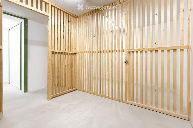 Empty hallway with white walls and a wooden partition featuring vertical slats. The floor is light-colored concrete, and there is a green door frame visible on the left side.