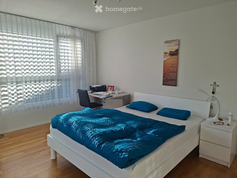 A well-lit bedroom with a large bed covered in a blue duvet, a nightstand with a lamp, a desk with a chair, and a printer. The room has wooden flooring and large windows with white curtains.
