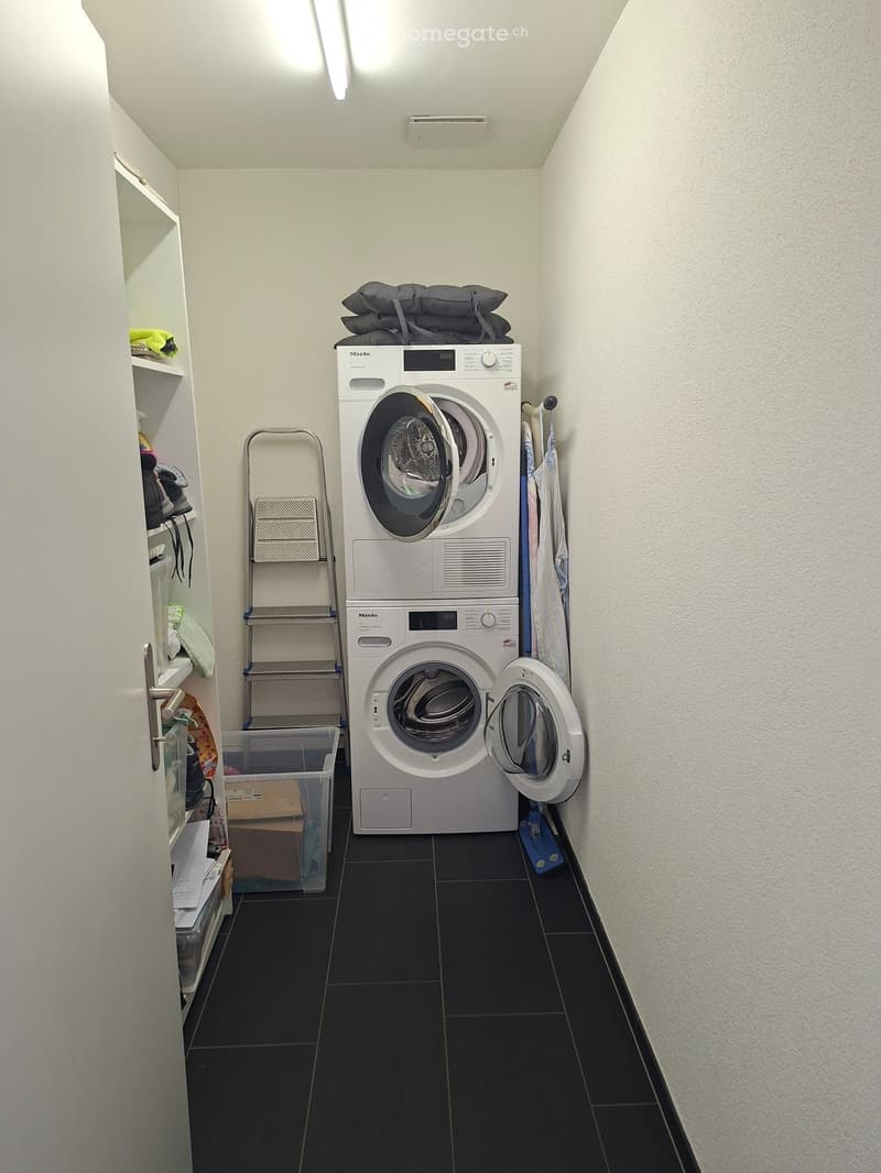A small, well-lit laundry area with a white washing machine, a metal ladder, a shelf with folded clothes, and a storage bin.