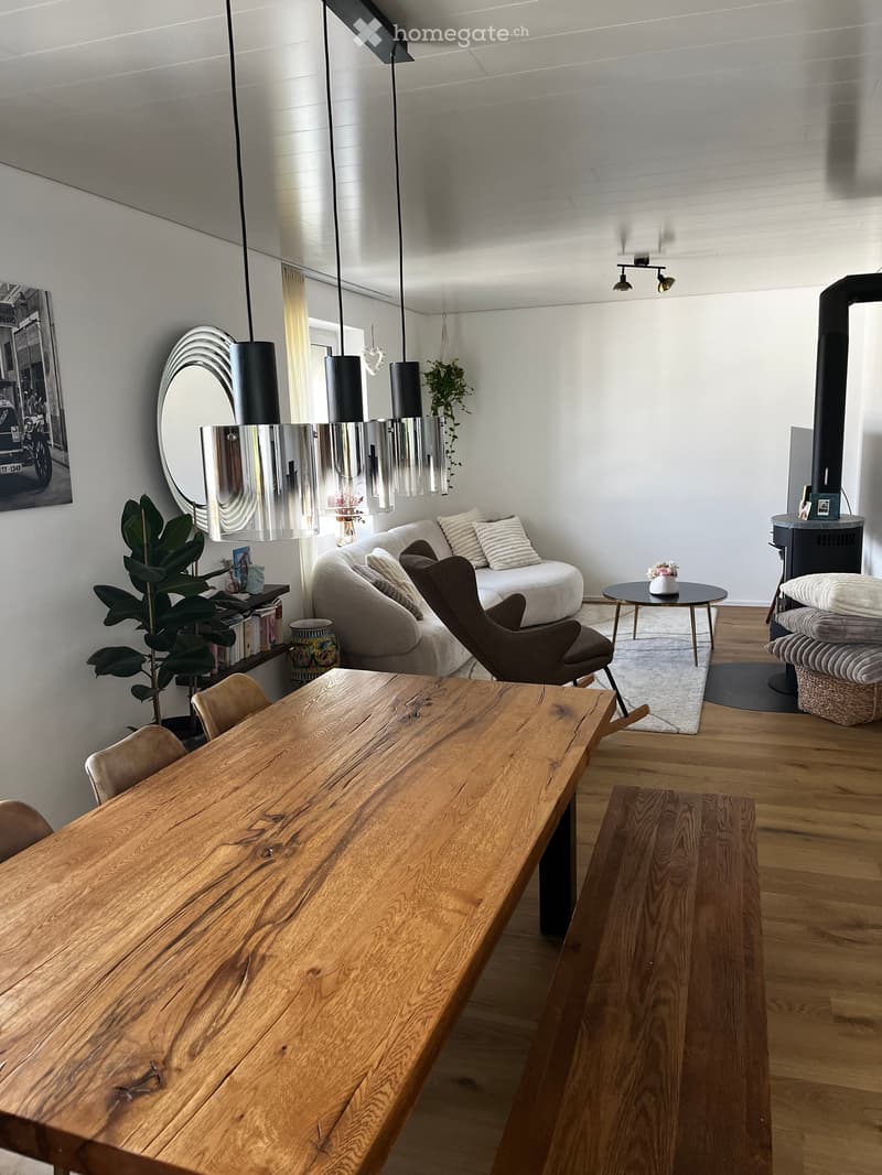Modern living room with a large wooden dining table, sofa, armchair, pendant lights, and a fireplace. The room features a mix of natural wood and neutral tones, with a plant and decorative items on a shelf.