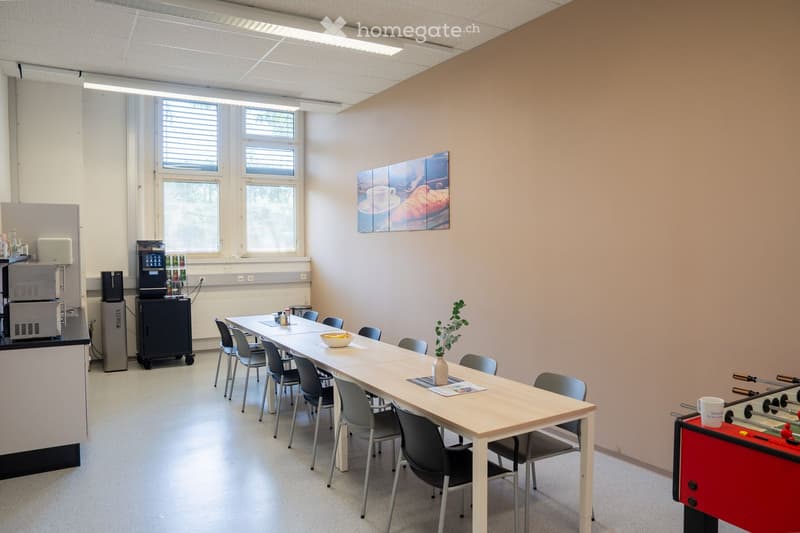Bright break room with a long wooden table surrounded by chairs, a coffee machine, refrigerator, a small plant, and a foosball table in the corner.
