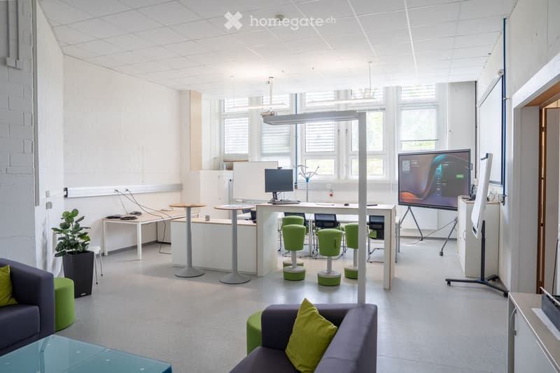 Modern office space with white brick walls, large windows, and a mix of seating and work areas. Features include a sofa, green stools, tables, chairs, a large screen, and a potted plant.