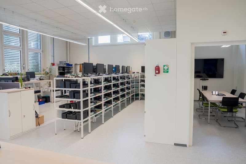 Modern office space with white walls, large windows, shelves filled with electronic components, a cabinet, a meeting table with chairs, and a large screen on the wall.