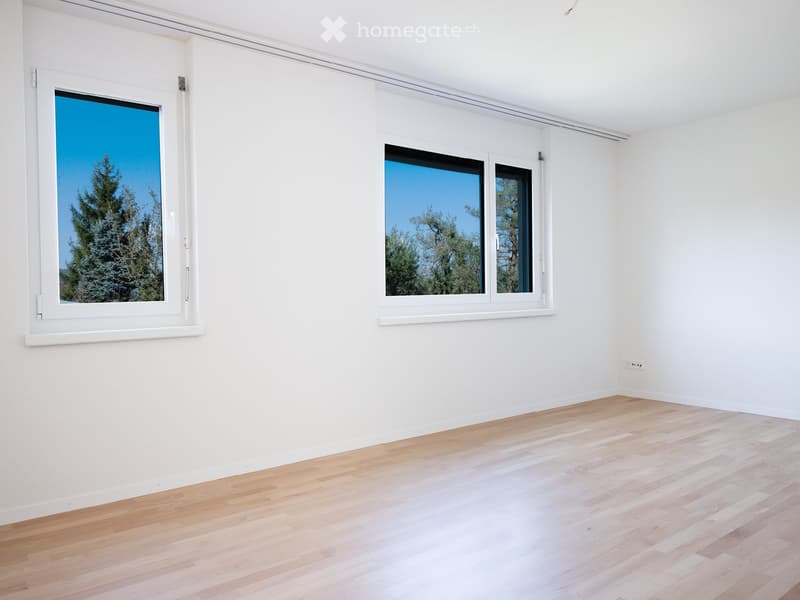 Empty room with white walls, light wooden flooring, and large windows showing a view of trees.