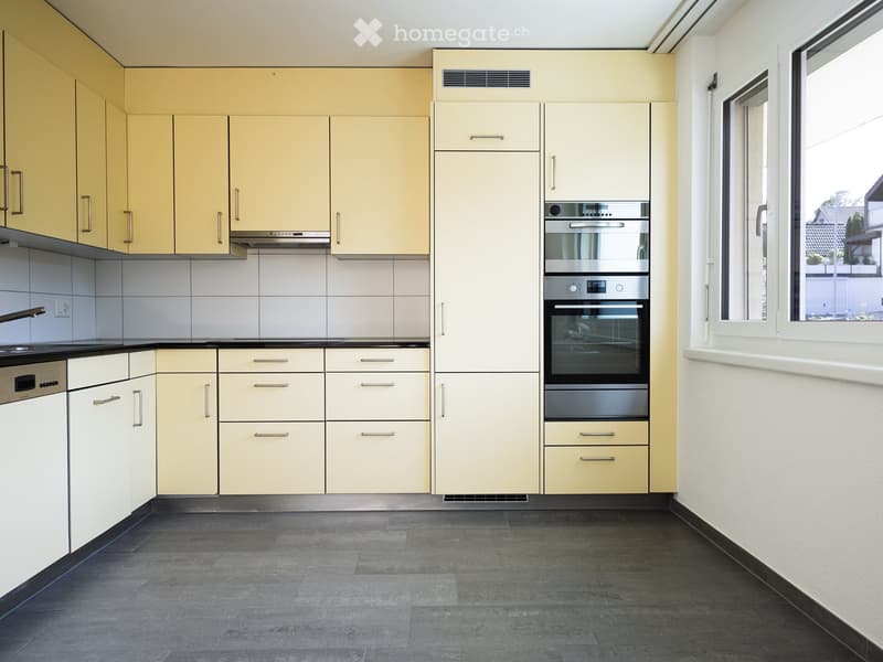 Modern kitchen with light-colored cabinets, stainless steel oven and dishwasher, dark tiled floor, and white tiled backsplash.
