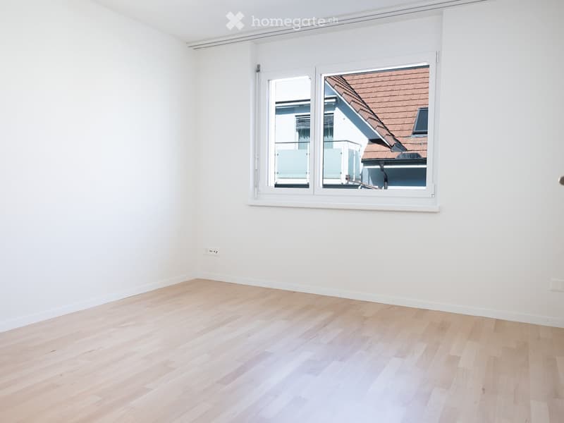 Empty room with light wooden flooring and white walls, featuring a large window with white frame showing a neighboring building with a red-tiled roof.