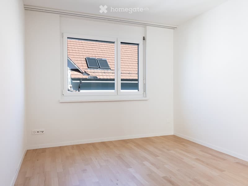 Empty room with light wooden flooring and white walls, featuring a large window with white frame showing a view of a tiled roof and part of a building.