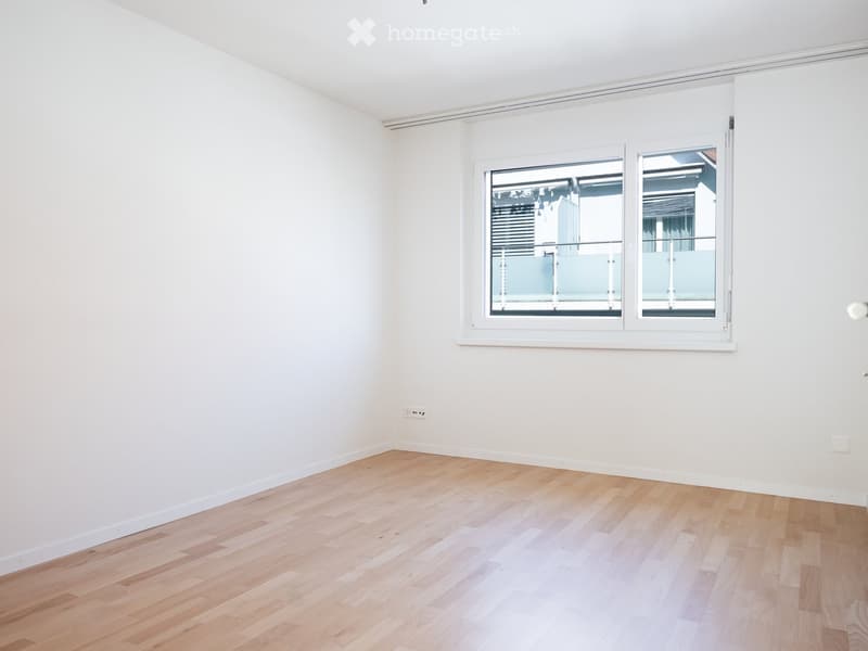 Empty room with light wooden flooring and white walls, featuring a large window with a view of a balcony area.