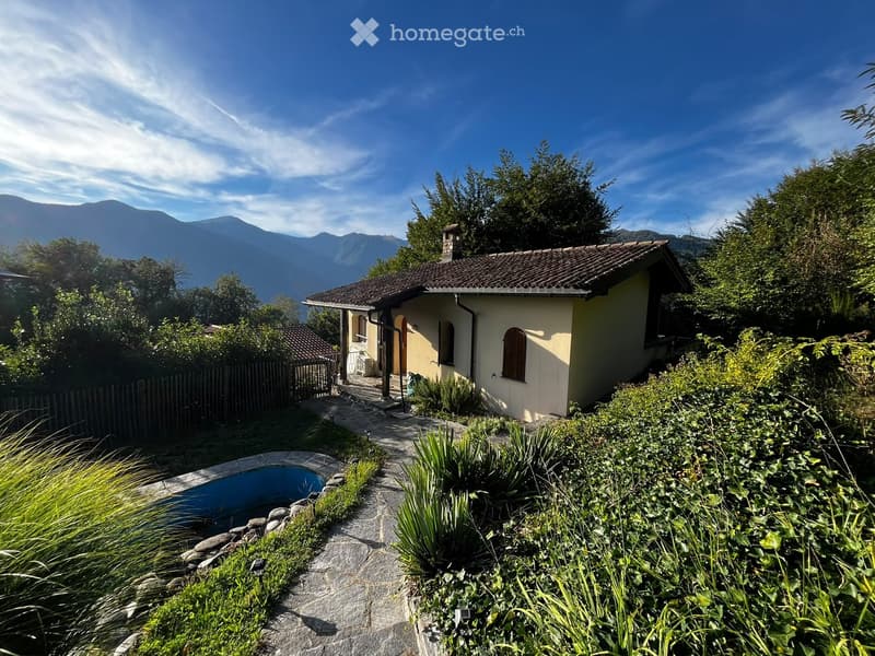 A small, single-story house with a tiled roof and light yellow walls, surrounded by a garden with a swimming pool, stone pathway, and lush greenery, with mountains visible in the background.