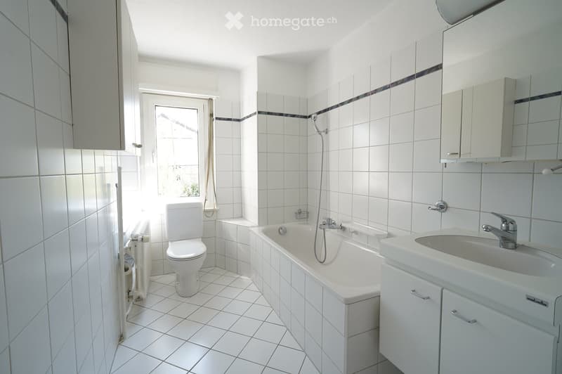 Bright bathroom with white tiled walls and floor, featuring a toilet, bathtub, sink, and mirrored cabinet.