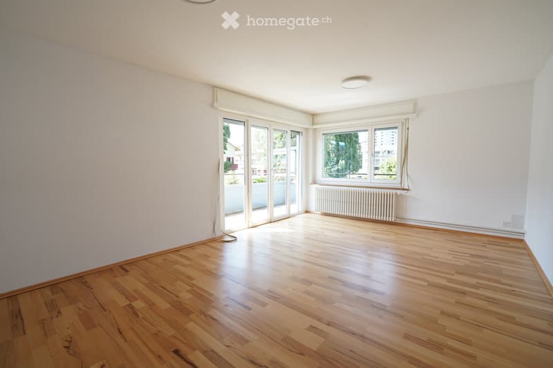 Empty room with wooden flooring, white walls, large windows, and a radiator. No furniture or equipment present.