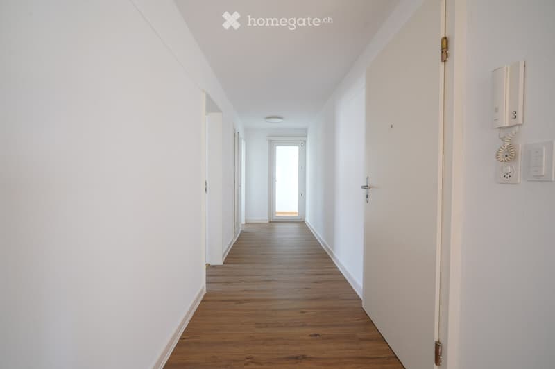 Bright, clean hallway with wooden flooring, white walls, and recessed lighting. Features a door at the end and a wall-mounted intercom system on the right side.