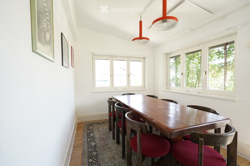 Bright dining room with a long wooden dining table surrounded by red-cushioned chairs, pendant lights, and framed artwork on the walls.