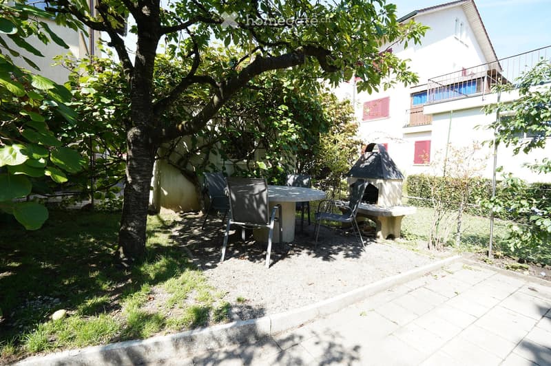 A small outdoor garden area with a round table, four chairs, a stone fireplace, and a large tree providing shade. The area is surrounded by a house with balconies and red shutters.