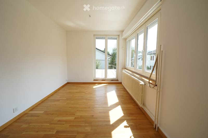 Empty room with wooden flooring, white walls, large windows, and a sliding glass door leading to a balcony.