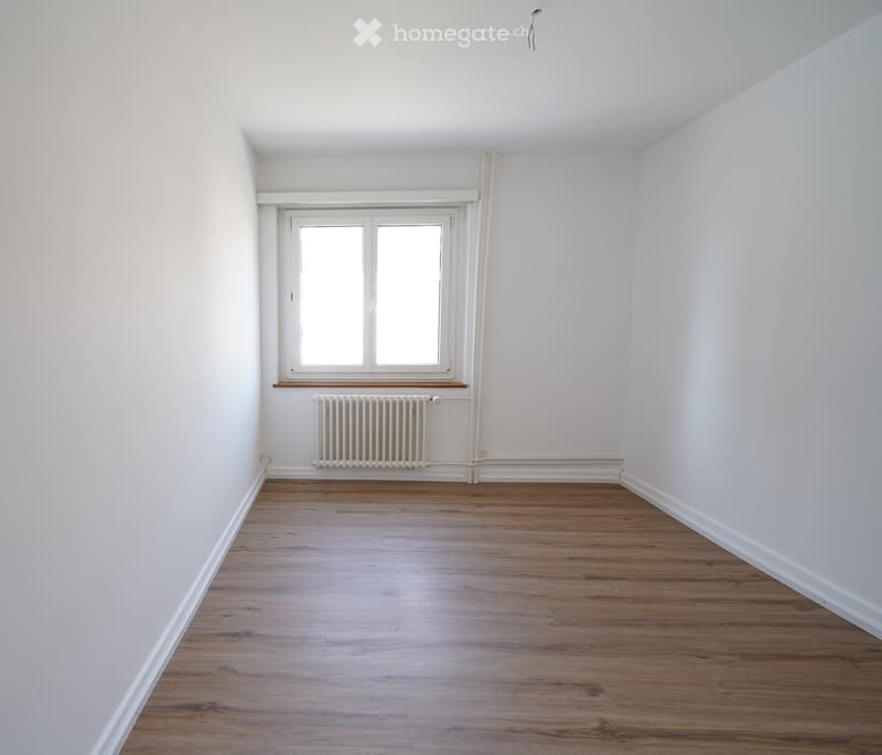 Empty room with light wooden flooring, white walls, a single window with white frame, and a radiator below the window.