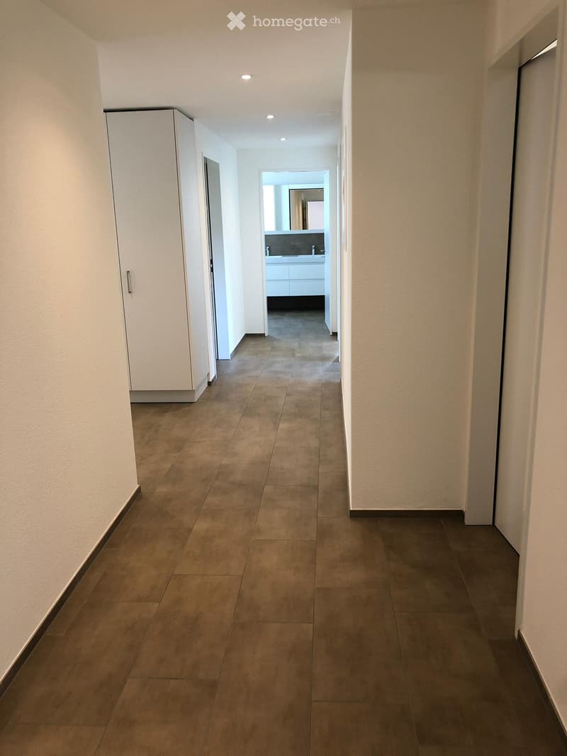 Clean, modern hallway with tiled flooring, white walls, and recessed lighting. A white cabinet is visible on the left, and a bathroom is visible at the end of the hallway.