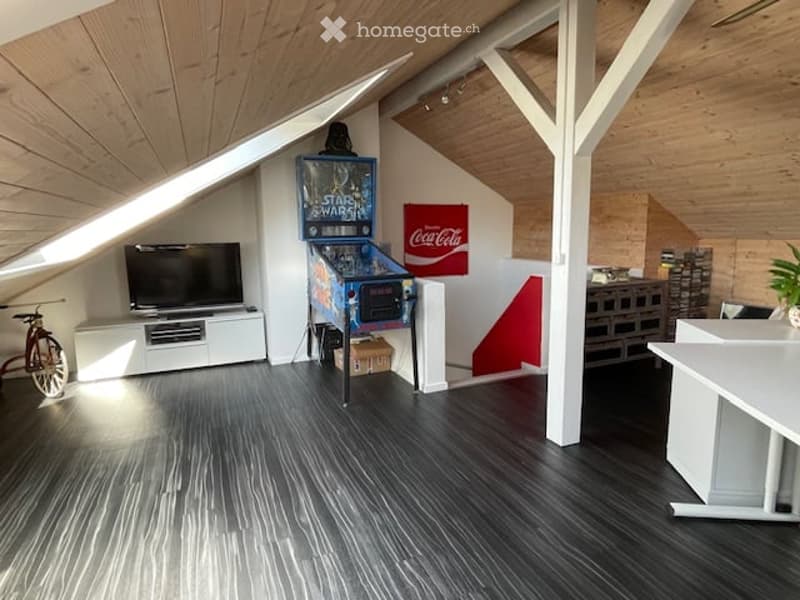Attic room with wooden ceiling beams, dark wood flooring, TV on a white stand, arcade game, bicycle, and a small table.
