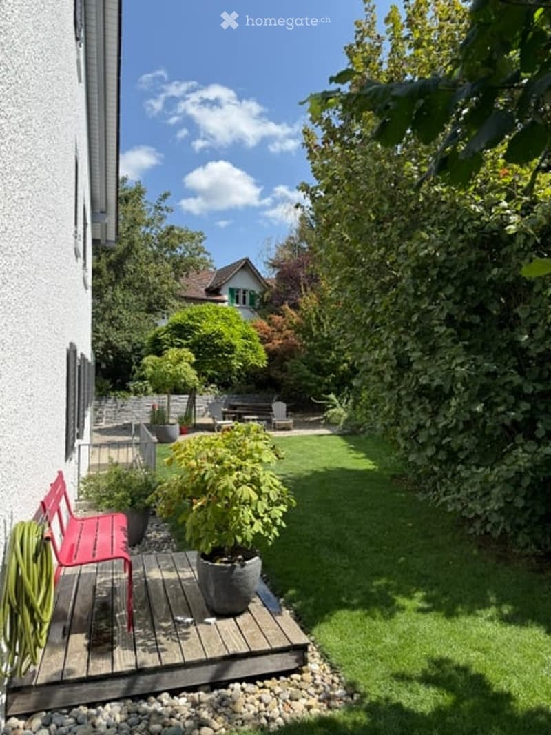 A well-maintained garden with a wooden deck, red bench, potted plants, and a grassy lawn, surrounded by trees and a neighboring house.