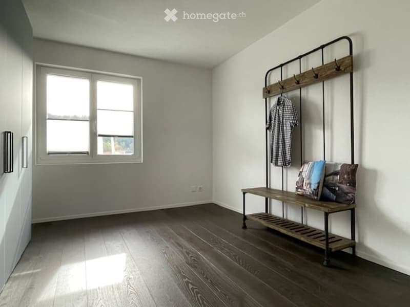 A modern, minimalist hallway with a wooden bench, coat rack, and picture frames. The room features light-colored walls, dark wooden flooring, and a large window allowing natural light to enter.
