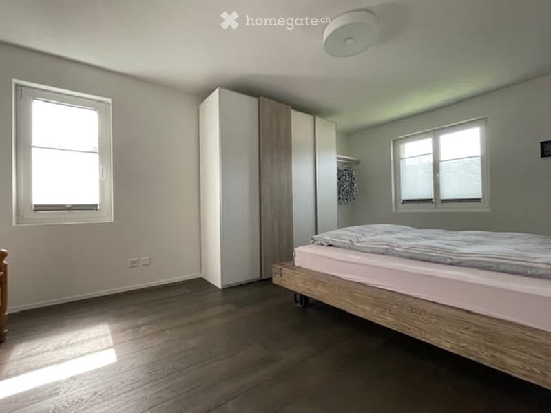 Bright bedroom with wooden flooring, a single bed, and a tall wardrobe. Two windows allow natural light to fill the space.