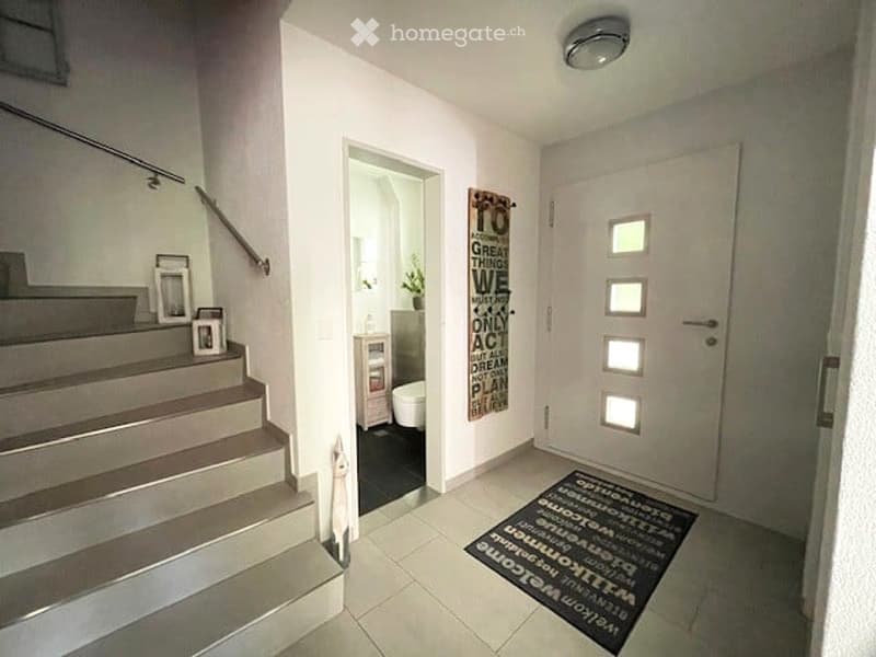 Modern hallway with white walls, tiled floor, staircase with metal railing, and a small decorative area with a plant and framed pictures.