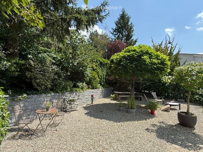 Well-maintained garden area with gravel ground, outdoor furniture including tables and chairs, potted plants, and trees providing shade.