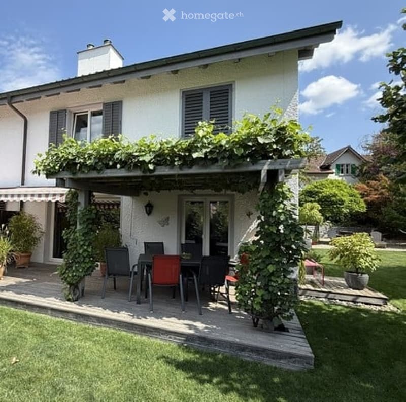 A covered patio area with a wooden deck, surrounded by greenery and climbing vines. A dining set with chairs and a table is placed on the deck.