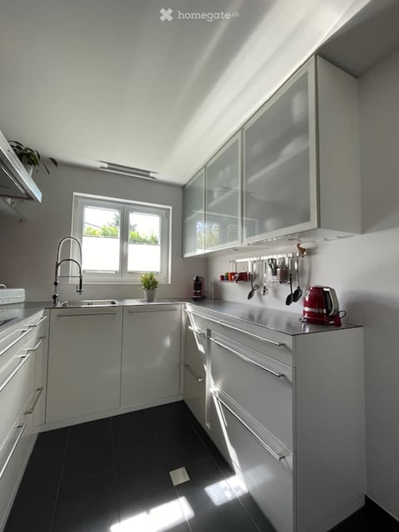 Modern, minimalist kitchen with white cabinets, black tiled floor, and stainless steel appliances. Features a central island with drawers, a sink with a modern faucet, and various kitchen utensils hanging on the wall.