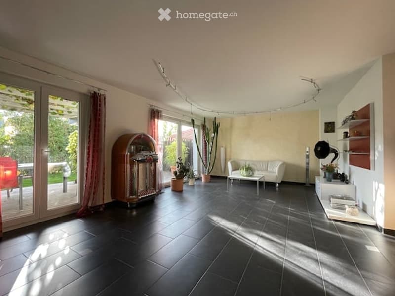 Bright living room with large windows and sliding glass doors leading to a patio. Features a white leather sofa, a coffee table, a vintage record player, several potted plants, and built-in shelves with decorative items.