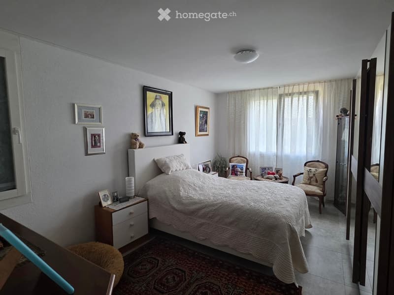 A well-lit bedroom with a large bed, nightstand, chair, small table, and several picture frames on the walls.