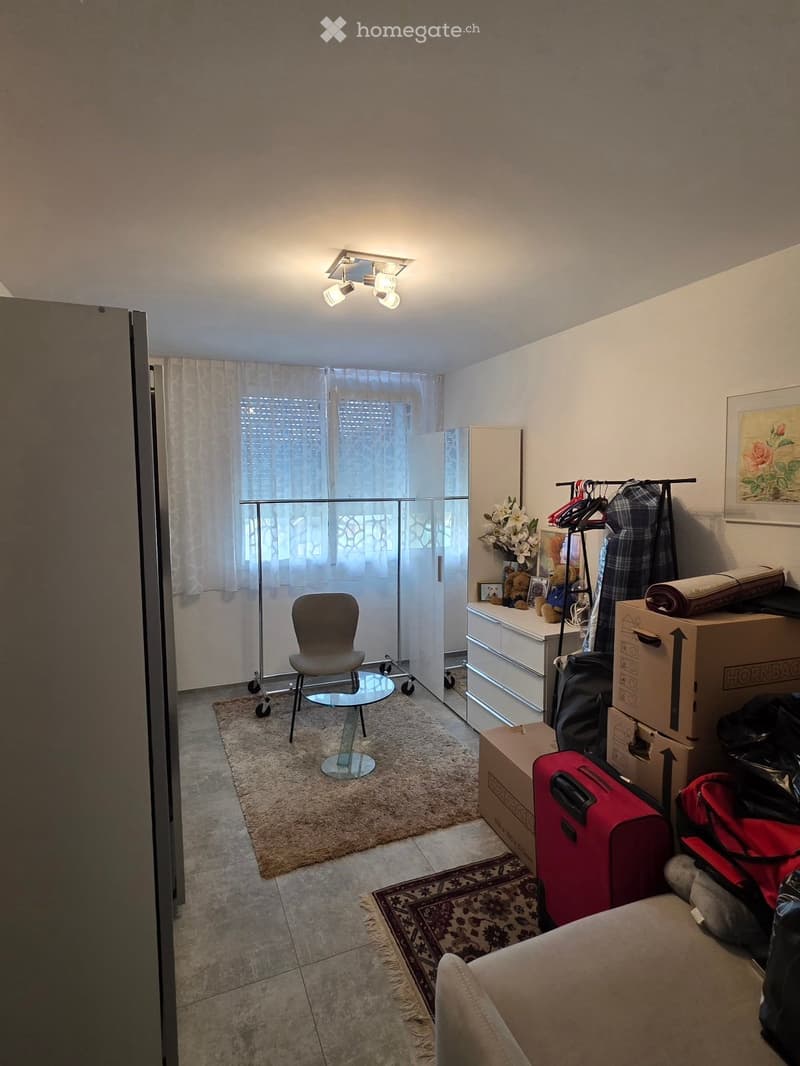 A furnished room with a chair, small table, sofa, dresser, suitcases, and a clothing rack. The room has tiled flooring, a carpet, and a window with curtains.