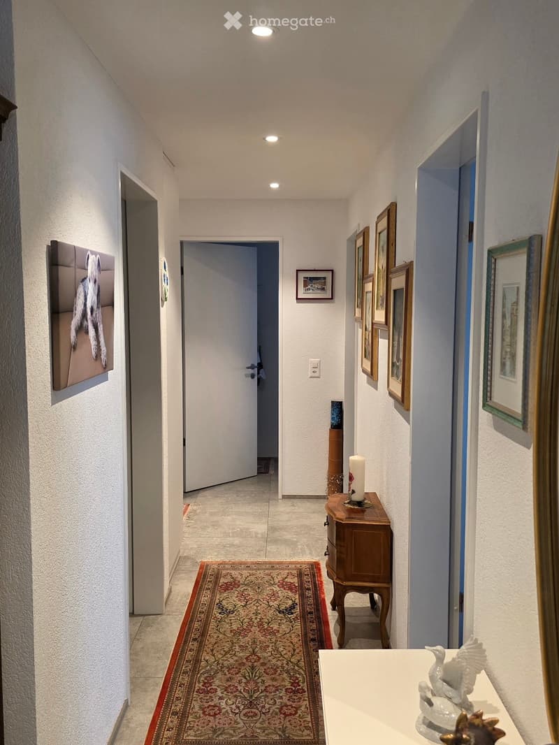 A well-lit hallway with white walls, a patterned rug, framed artwork, and a small wooden table with a candle and decorative items.