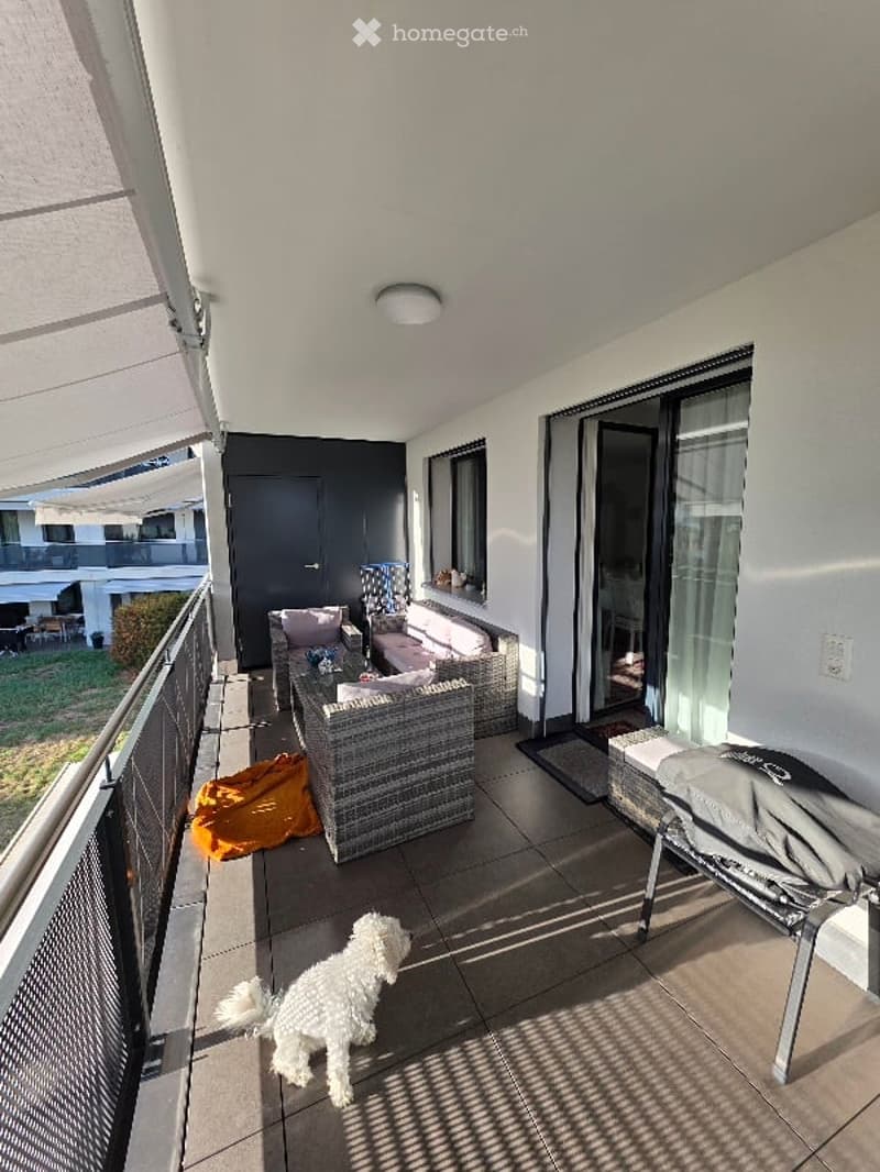 Balcony with wicker seating, small table, and a white dog. Features large windows, a sliding glass door, and a covered area with a canopy.