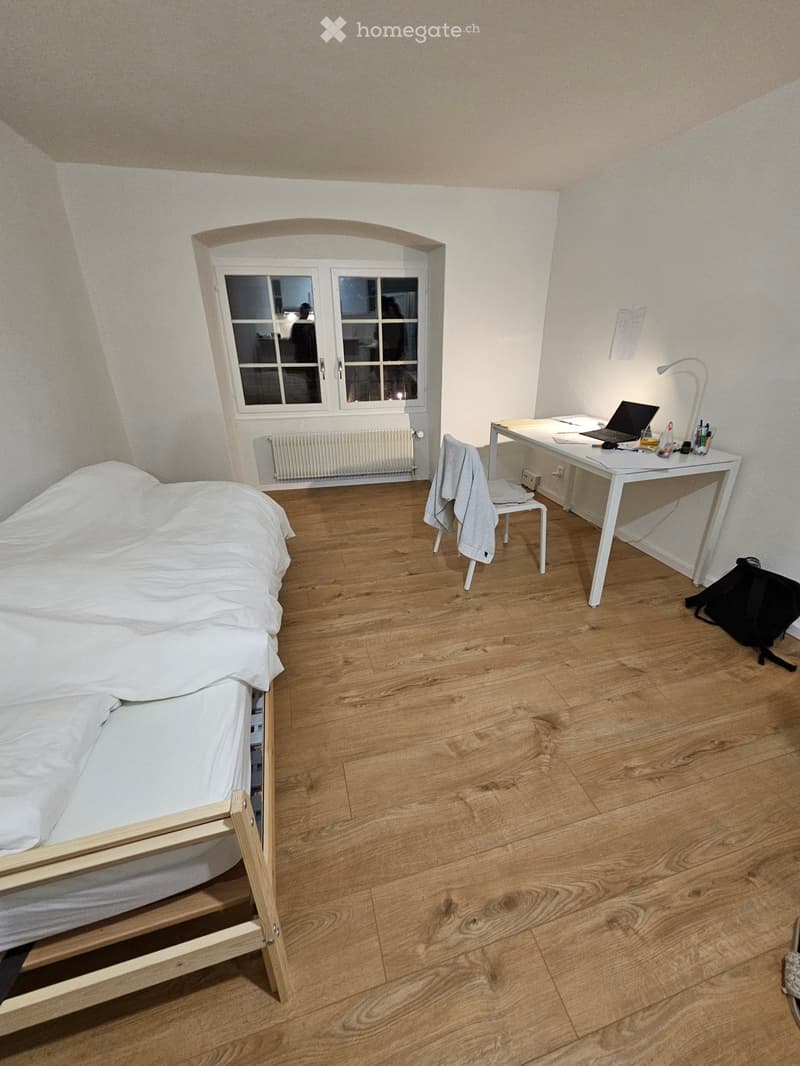 Cozy bedroom with wooden floor, single bed, desk with laptop, chair, and a backpack on the floor.