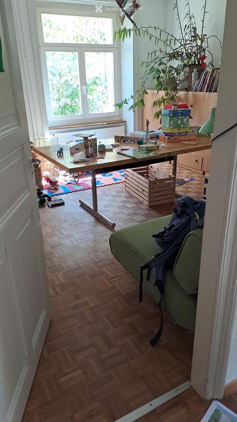A well-lit living room with wooden flooring, a green sofa, a large wooden table, and a plant in the corner. The room features a mix of furniture and personal items, including a backpack on the sofa and books on the table.