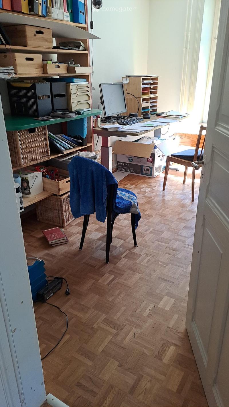 A well-lit study or office space with wooden flooring, a desk with a computer setup, shelves filled with books and office supplies, and a comfortable chair.