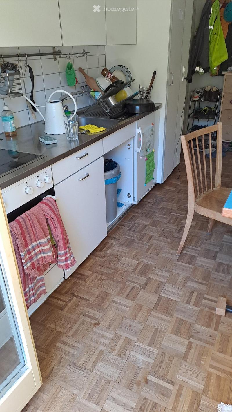 The kitchen features white cabinets, a wooden herringbone floor, and a stainless steel sink with a dish rack. A small table and chair are visible in the foreground, and various kitchen items such as oven mitts, dish soap, and cleaning supplies are scatter