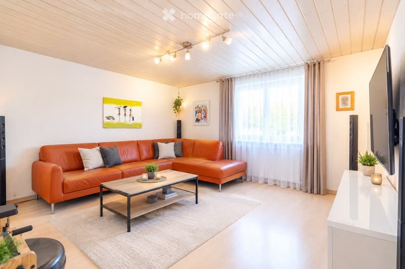 Modern living room with an orange leather sofa, coffee table, and large windows with curtains. Features include a TV, speakers, and decorative plants.