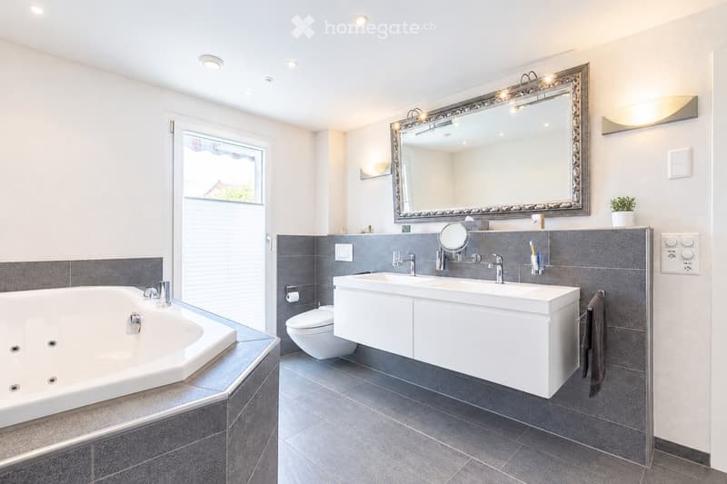 Modern bathroom with a large bathtub, double sink vanity, toilet, and a large ornate mirror above the sink. The walls are tiled in grey, and the floor is covered with dark grey tiles. A small window provides natural light, and a towel hangs on a rack.
