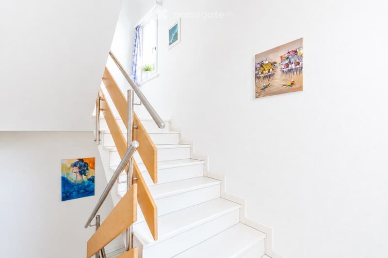 Modern staircase with white steps, wooden handrails, and metal balusters. Two abstract paintings are displayed on the walls, and a small potted plant sits on a windowsill.