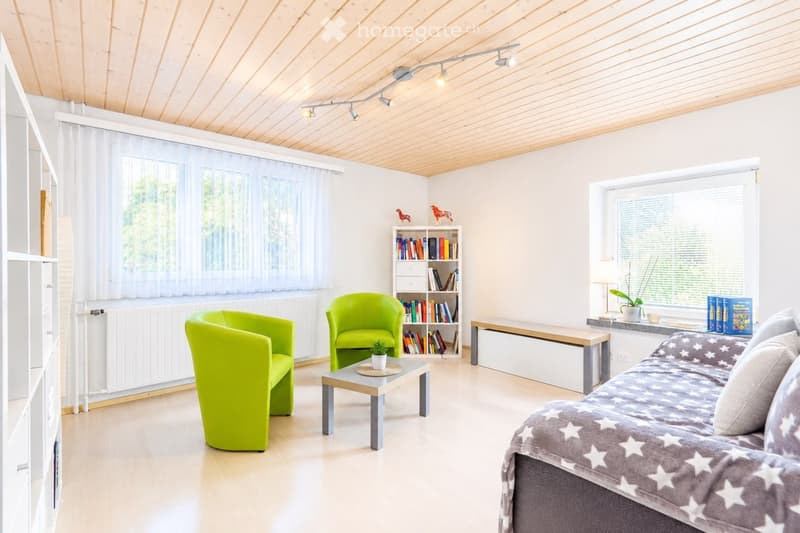 Bright living room with wooden floors, white walls, and large windows. Features a grey sofa with a star-patterned throw, two green armchairs, a small coffee table, a bookshelf, and a bench.