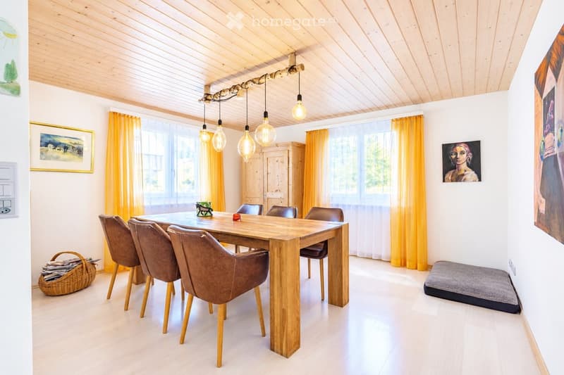 Bright dining area with a wooden dining table surrounded by brown leather chairs, a wicker basket on the floor, and large windows with yellow curtains. The room features a wooden ceiling with exposed beams and pendant lights.