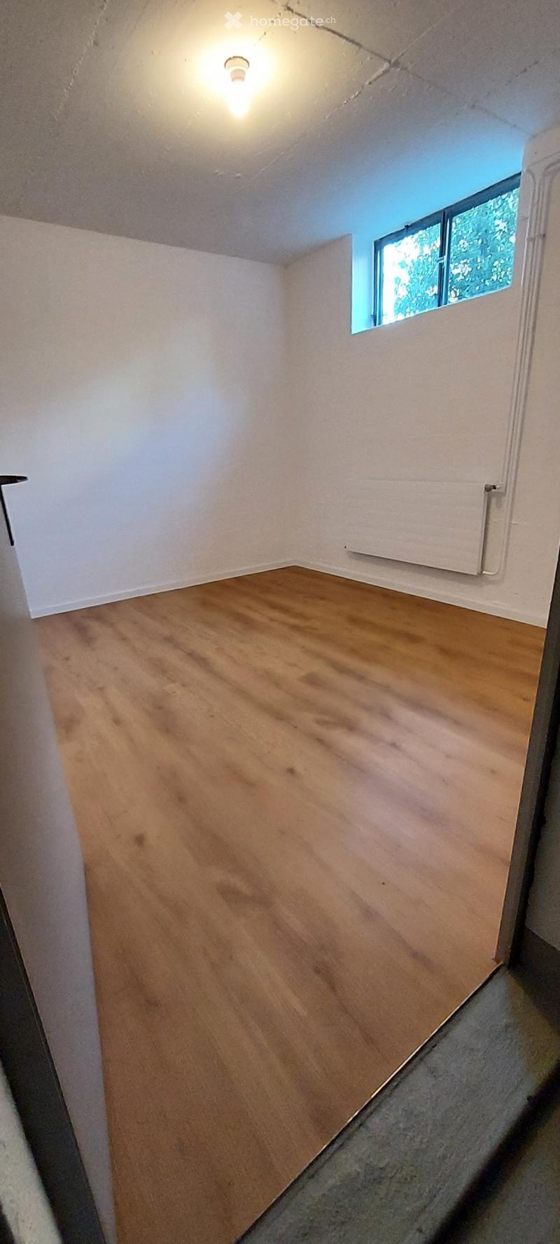 Empty room with wooden flooring, white walls, and a small window letting in natural light.
