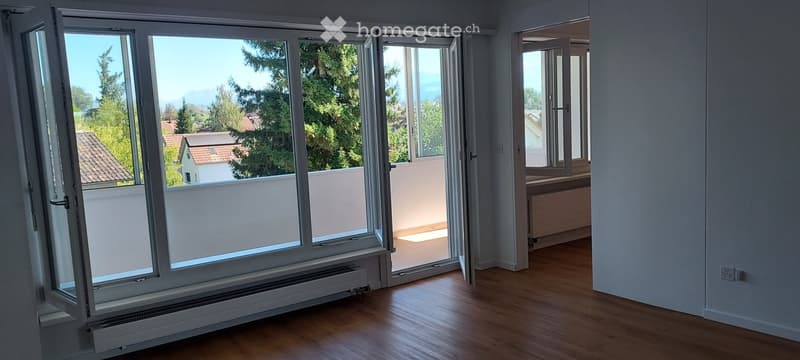 Empty room with wooden flooring, large windows, and a view of the balcony and surrounding greenery.