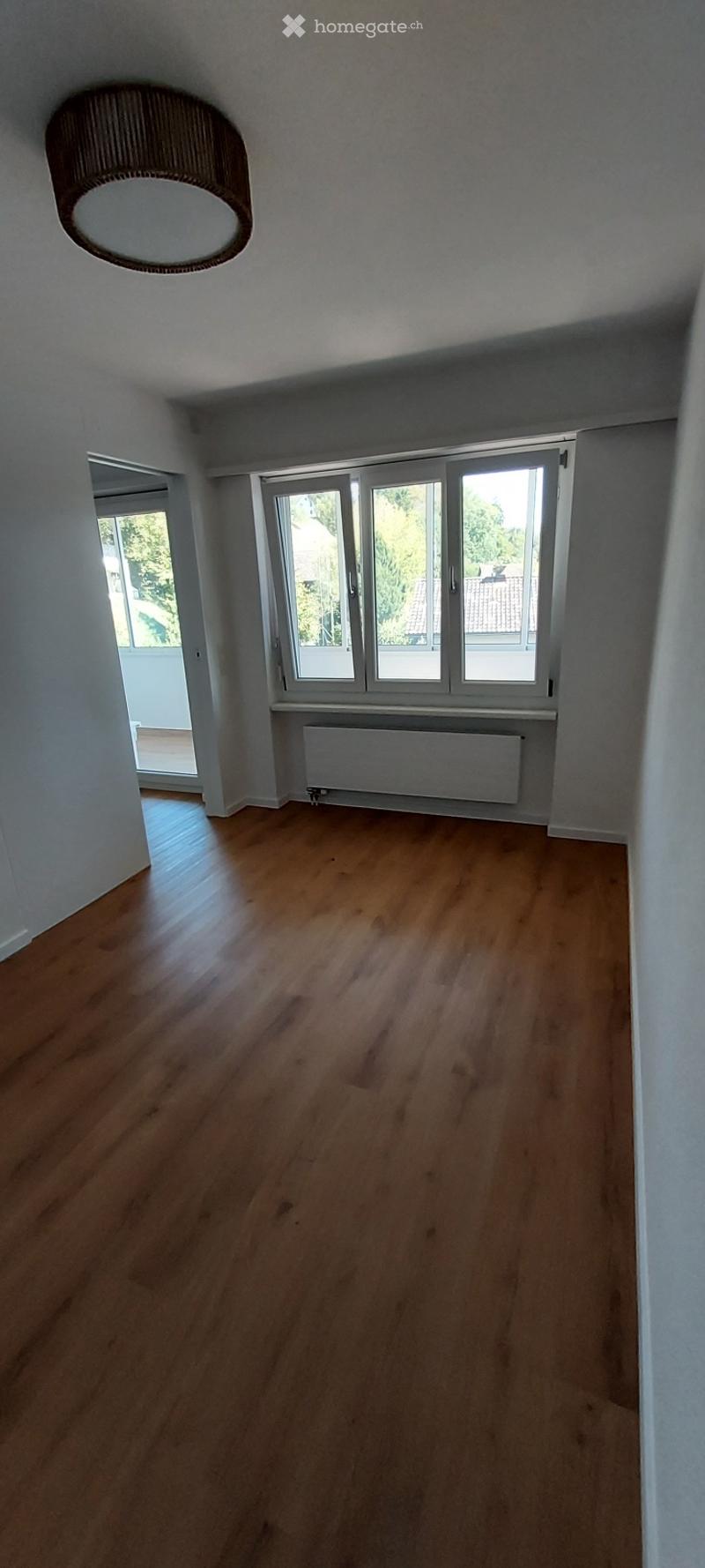 Empty room with wooden flooring, white walls, large windows, and a visible radiator. No furniture or equipment present.